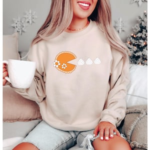 Puede incluir: Sudadera beige con un gráfico de tarta retro, con una porción faltante, flores de margarita y tres montones de crema batida. Esta parte superior de manga larga es una prenda informal, perfecta para el uso diario.