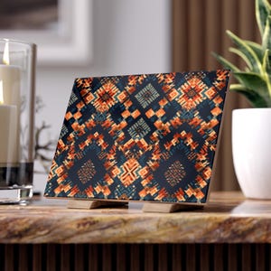 May include: A decorative square tile with a geometric pattern in shades of navy blue, orange, and teal. The tile is propped up on two small wooden stands. The tile is displayed on a marble surface.