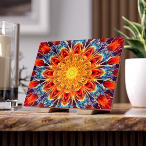 May include: A square decorative art piece with a vibrant, symmetrical floral design in shades of orange, yellow, blue, and purple. The artwork is displayed on a wooden stand, showcasing a kaleidoscope-like pattern. The piece is approximately 6 inches square.