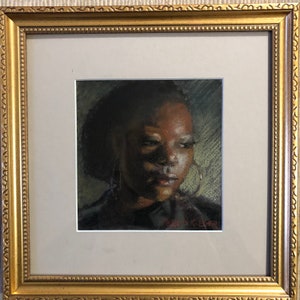 Op de afbeelding: Een ingelijste pasteltekening van een persoon met een donkere huid en kort donker haar. De persoon draagt grote oorringen en kijkt naar beneden en naar links. De tekening is in een gouden lijst.