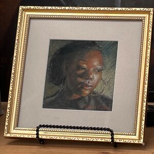 Op de afbeelding: Een ingelijst portret van een persoon met een donkere huidskleur, die een donker shirt en grote creolen draagt. Het portret is in een gouden lijst met een decoratieve rand.