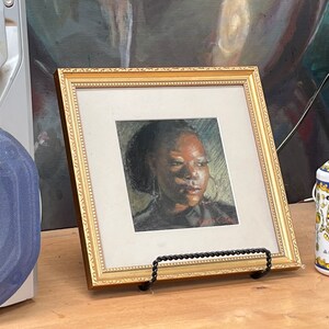 Op de afbeelding: Een ingelijst portret van een vrouw met een donkere huid en bruine ogen. Het portret is in een gouden lijst en staat op een houten tafel. Rechts van het portret staat een fles handzeep met een bloemenmotief.