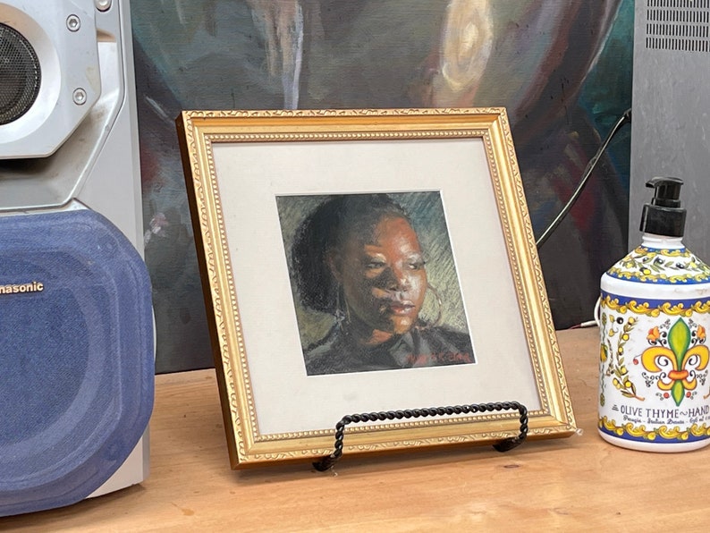 Op de afbeelding: Een ingelijst portret van een vrouw met een donkere huid en bruine ogen. Het portret is in een gouden lijst en staat op een houten tafel. Rechts van het portret staat een fles handzeep met een bloemenmotief.
