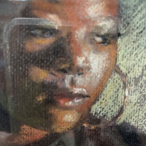 Op de afbeelding: Een close-up portret van een persoon met een donkere huid, bruine ogen en een neutrale uitdrukking. De persoon draagt een donker shirt en grote creolen. Het portret is getekend in pastelkleuren op een donkere achtergrond. De handtekening van de kunstenaar, "Marilyn Reger", is zichtbaar in de rechterbenedenhoek.