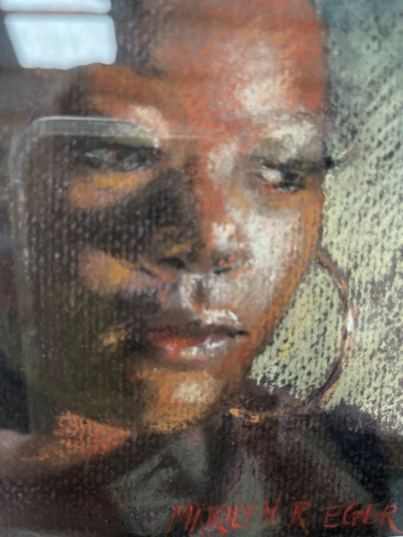 Op de afbeelding: Een close-up portret van een persoon met een donkere huid, bruine ogen en een neutrale uitdrukking. De persoon draagt een donker shirt en grote creolen. Het portret is getekend in pastelkleuren op een donkere achtergrond. De handtekening van de kunstenaar, "Marilyn Reger", is zichtbaar in de rechterbenedenhoek.