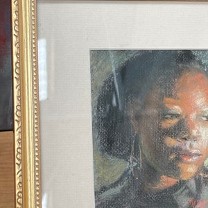 Op de afbeelding: Een ingelijste pasteltekening van een gezicht. De persoon heeft donker haar en draagt een donker shirt. De tekening is in een gouden lijst.
