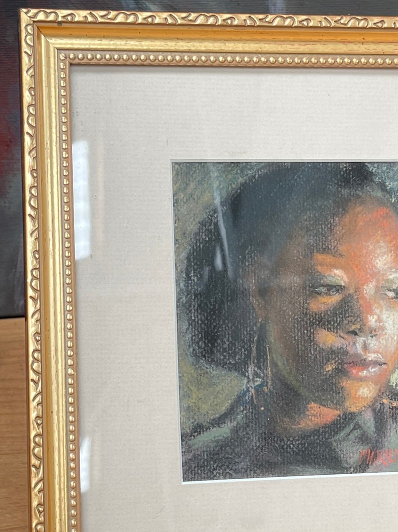 Op de afbeelding: Een ingelijste pasteltekening van een gezicht. De persoon heeft donker haar en draagt een donker shirt. De tekening is in een gouden lijst.