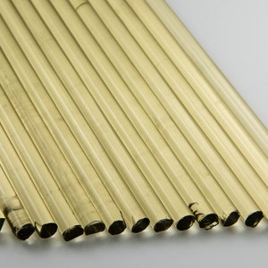 Könnte beinhalten: Eine Ansammlung klarer, zylindrischer Glasröhren, die diagonal auf einer weißen Oberfläche angeordnet sind. Die Röhren haben einen leichten Bernsteinton und sind gleichmäßig angeordnet, wodurch ein Muster aus parallelen Linien entsteht.