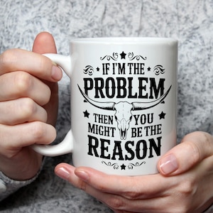 Puede incluir: Taza de cerámica blanca con un gráfico negro que dice "IF I'M THE PROBLEM THEN YOU MIGHT BE THE REASON" con un diseño de calavera de toro. La taza tiene un asa curva y está sostenida por una persona.