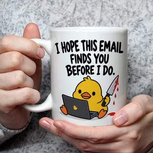 Può includere: Una tazza in ceramica bianca con un'anatra gialla dei cartoni animati seduta davanti a un computer portatile e che tiene in mano un coltello con gocce di sangue rosso. Il testo sopra la grafica recita, I HOPE THIS EMAIL FINDS YOU BEFORE I DO.