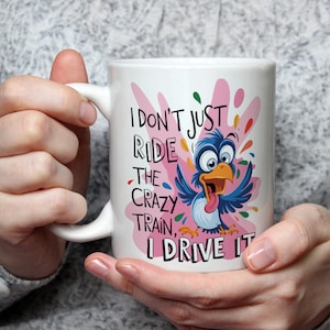 May include: White ceramic mug featuring a cartoon bird illustration and the text "I don't just ride the crazy train, I drive it." The bird is blue with an orange beak and tongue, set against a pink and colourful splatter design.