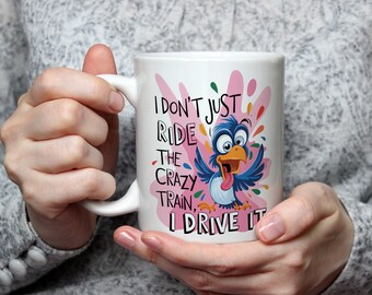 May include: White ceramic mug with a cartoon bird illustration and the text "I don't just ride the crazy train, I drive it." The bird is blue with an orange beak and tongue, set against a pink and colorful splatter design.