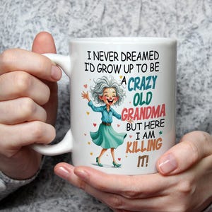 May include: White ceramic mug with a handle featuring a cartoon illustration of a smiling elderly woman in a teal dress. The mug has the text: "I NEVER DREAMED I'D GROW UP TO BE A CRAZY OLD GRANDMA BUT HERE I AM KILLING IT!"