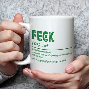 May include: White ceramic mug with a green shamrock and the word "FECK" in green letters. The definition of the word is written below in black text: "(fek) verb Ireland's most used word. A uniquely Irish way of expressing one's annoyance or frustration. See also feck off feck sake, feckin eejit"