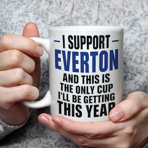 May include: A white ceramic mug featuring the blue and black text I SUPPORT EVERTON AND THIS IS THE ONLY CUP I'LL BE GETTING THIS YEAR. The mug has a curved handle on the left side and a smooth, cylindrical body with a clean, simple design.