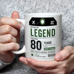 May include: A white ceramic mug featuring a graphic design resembling a luggage tag. The text reads DESTINATION LEGEND, PASSENGER 80 YEARS, STATUS FIRST CLASS, PRIORITY BOARDING, and BAGGAGE: 80 YEARS OF STORIES. An airplane icon and barcodes are included.