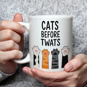 May include: White ceramic mug with the text "CATS BEFORE TWATS" in black. The mug features four colorful cat paw prints below the text. The paws are in orange, gray, black and white with pink pads.