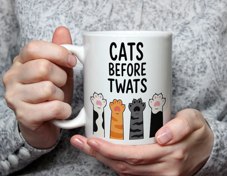 May include: White ceramic mug with the text "CATS BEFORE TWATS" in black. The mug features four colorful cat paw prints below the text. The paws are in orange, gray, black and white with pink pads.