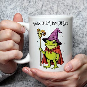 May include: White ceramic mug with a cartoon frog wizard, wearing a purple hat and red cape, holding a staff with a skull. The mug has the text "Twas the Tism M'Lord".
