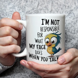 Könnte beinhalten: Weiße Keramik-Tasse mit dem humorvollen Zitat: "I'M NOT RESPONSIBLE FOR WHAT MY FACE DOES WHEN YOU TALK." Die Tasse zeigt einen Cartoon-Vogel mit großen Augen und blauen, gelben und grünen Federn. Die Tasse wird von einer Person gehalten.