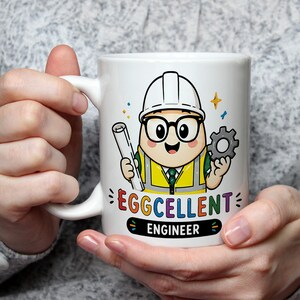 Könnte beinhalten: Ein weißer Keramikbecher mit einem Cartoon-Ei-Charakter, der einen weißen Schutzhelm, eine Brille und eine gelbe Warnweste trägt. Die Figur hält eine aufgerollte Blaupause und ein graues Zahnrad. Darunter steht der Text EGGCELLENT ENGINEER.
