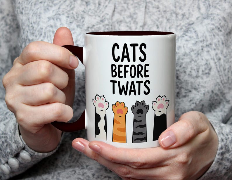 May include: White ceramic mug with a dark brown interior and handle. The mug features the text "CATS BEFORE TWATS" in black, above illustrations of colorful cat paws. The paws are black and white, orange, gray, and pink.