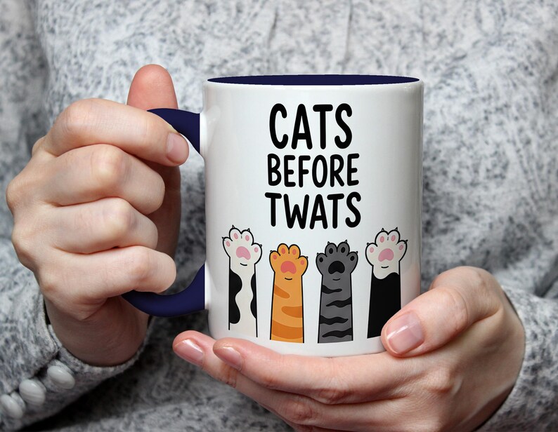 May include: White ceramic mug with a navy blue handle and interior. The mug features the text "CATS BEFORE TWATS" in black, above a row of colorful cat paw prints. The mug is held by a person.