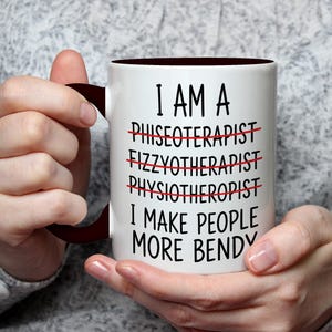 May include: White ceramic mug with a dark brown interior and handle. The mug features the text "I AM A... I MAKE PEOPLE MORE BENDY" with crossed-out words. The mug is held by a person.