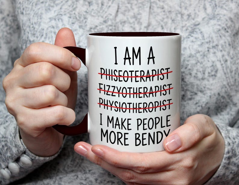 May include: White ceramic mug with a dark brown interior and handle. The mug features the text "I AM A... I MAKE PEOPLE MORE BENDY" with crossed-out words. The mug is held by a person.