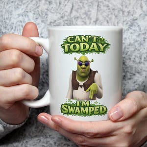 May include: A white ceramic mug featuring a graphic of a green ogre wearing dark sunglasses, a brown vest, and a white shirt. The text above the character reads CAN'T TODAY and the text below reads I'M SWAMPED, both set against a green slime background.