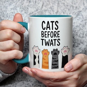 May include: White ceramic mug with a teal handle and interior. The mug features the text "CATS BEFORE TWATS" in black, with illustrations of cat paws in various colors below the text. A humorous cat-themed gift.