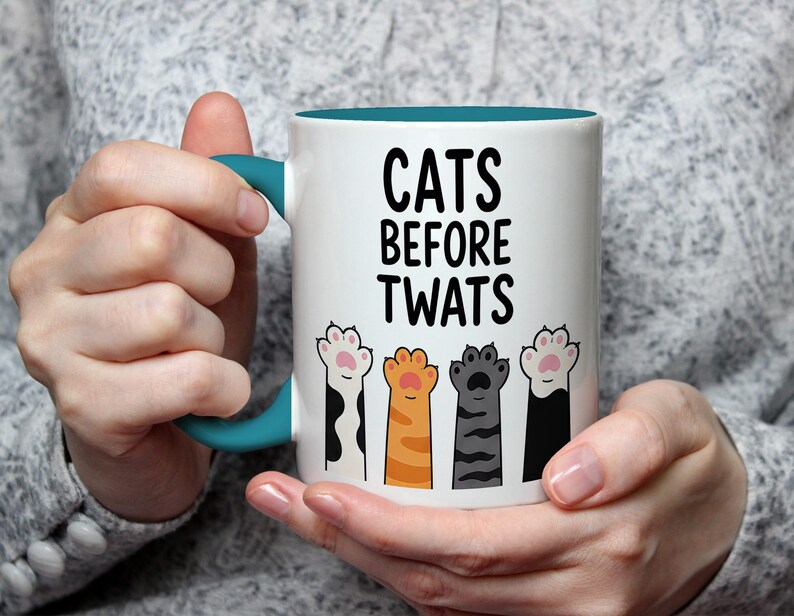 May include: White ceramic mug with a teal handle and interior. The mug features the text "CATS BEFORE TWATS" in black, with illustrations of cat paws in various colors below the text. A humorous cat-themed gift.