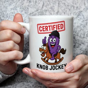 May include: A white ceramic mug featuring a cartoon graphic of a purple creature wearing a black helmet, riding a brown rocking horse. Above the graphic is a red rectangular stamp with the text CERTIFIED, and below is the text KNOB JOCKEY.