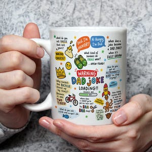 Peut inclure: Un mug en céramique blanche orné d'illustrations colorées et de textes tels que 'WARNING DAD JOKE LOADING!!', 'NACHO CHEESE!', 'KING OF PUNS', ainsi que diverses blagues sur les grenouilles, vélos, moutons et pieuvres avec une barre de progression.