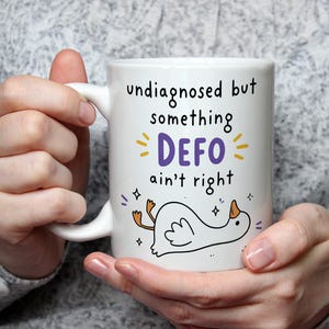 May include: White ceramic mug with the text "undiagnosed but something DEFO ain't right" in black and purple. A cartoon duck is drawn below the text. The mug is held by a person.