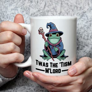 Può includere: Tazza in ceramica bianca con un disegno di rana mago dei cartoni animati. La rana indossa una veste e un cappello blu, con un bastone con un teschio. La tazza ha il testo "Twas the 'tism M'Lord" stampato.