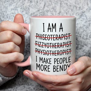 May include: White ceramic mug with a pink interior and handle. The mug has the text "I AM A... I MAKE PEOPLE MORE BENDY" with crossed-out words. The mug is held by a person.