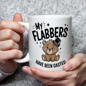 以下が含まれることがあります： 「My Flabbers Have Been Gasted」の文字とシルクハットをかぶったクマの漫画が描かれた白いセラミックマグ。クマは悲しそうな表情をしています。マグカップは人が持っています。