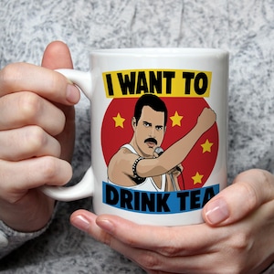 May include: White ceramic mug with a cartoon illustration of a man with a mustache holding a microphone. The design is set against a red circle with yellow stars and the text "I WANT TO DRINK TEA" in yellow and blue.