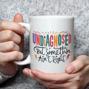 May include: A white ceramic mug with the words "UNDIAGNOSED But somethin Ain't Right" in colourful lettering. The mug is held by a person. The mug has a white handle.