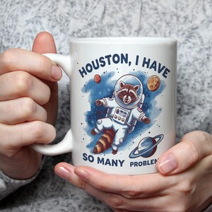 Puede incluir: Taza de cerámica blanca con un dibujo animado de un mapache con un traje espacial y el texto "Houston, tengo tantos problemas".