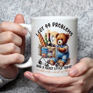 May include: White ceramic mug featuring a brown teddy bear pushing a shopping cart filled with groceries. The mug has the text "I got 99 problems and a sweet little treat would solve most of the".