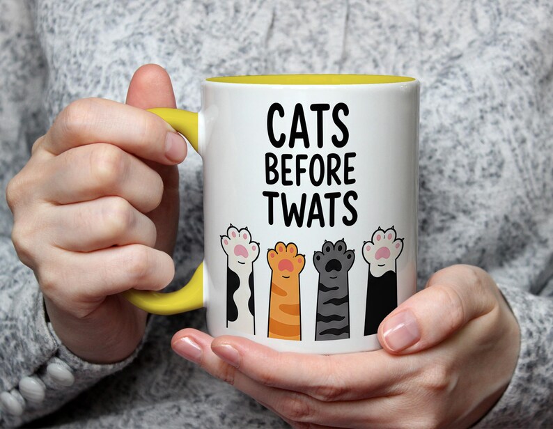 May include: White ceramic mug with a yellow handle and interior, featuring the text "CATS BEFORE TWATS" in black. Below the text are four colorful cat paw prints. The mug is held by a person.