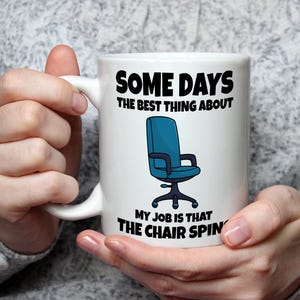 May include: White ceramic mug with the text "SOME DAYS THE BEST THING ABOUT MY JOB IS THAT THE CHAIR SPINS". The mug features a blue office chair graphic. The mug is held by a person wearing a grey sweater.