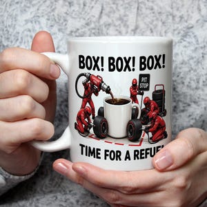 May include: A white ceramic mug featuring a graphic of a Formula 1 pit crew in red uniforms servicing a smaller coffee mug. The text reads BOX! BOX! BOX! and TIME FOR A REFUEL. The design includes a fuel hose, tyres, and a pit stop sign.
