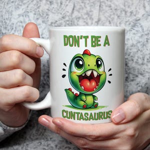 May include: White ceramic mug with a cartoon dinosaur design and the text "DON'T BE A CUNTASAURUS". The dinosaur is green with a red crest and open mouth. The mug is held by a person wearing a grey jumper.