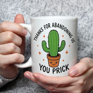 May include: White ceramic mug featuring a green cactus in a brown pot with a smiling face. The mug has the text "THANKS FOR ABANDONING US YOU PRICK" in black, with orange star accents. The mug is held by a person.