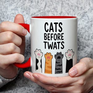 May include: White ceramic mug with a red interior and handle, featuring the text "CATS BEFORE TWATS" in black. The mug is decorated with colorful cat paw prints. The mug is held by a person.