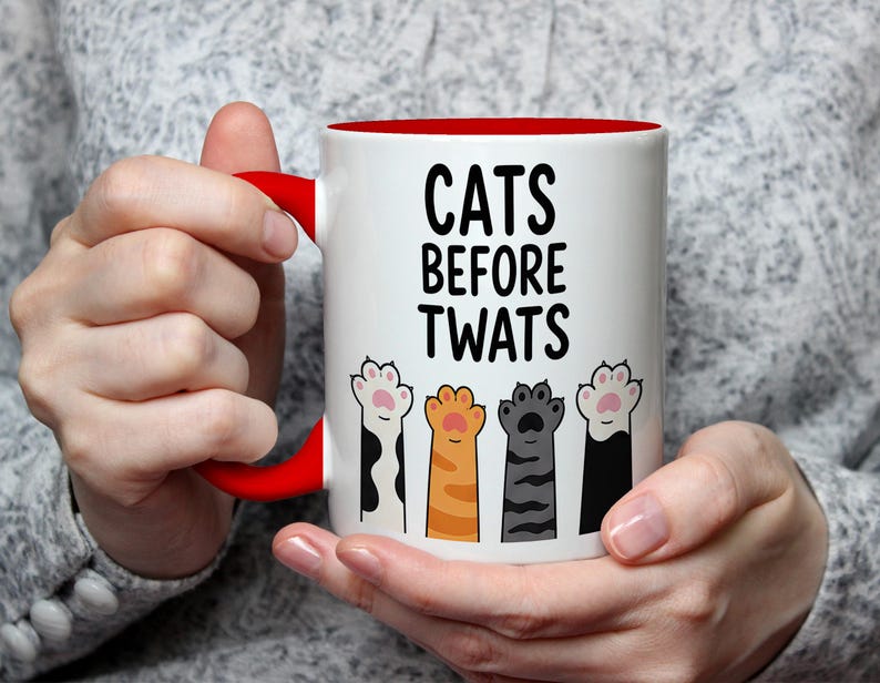 May include: White ceramic mug with a red interior and handle, featuring the text "CATS BEFORE TWATS" in black. The mug is decorated with colorful cat paw prints. The mug is held by a person.