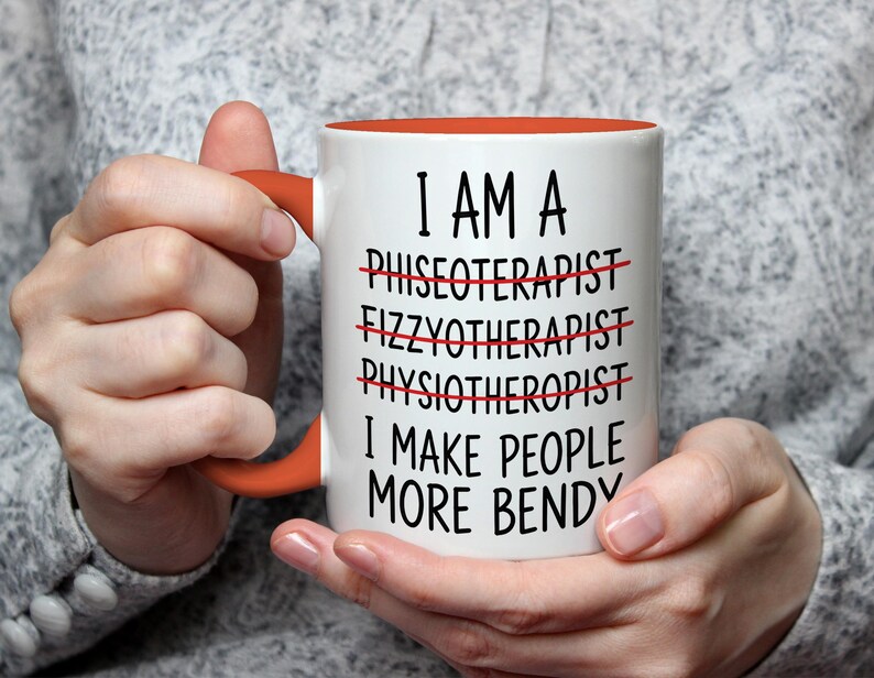 May include: White ceramic mug with an orange interior and handle. The mug features the text "I AM A... I MAKE PEOPLE MORE BENDY" with crossed-out words. The mug is held by a person.